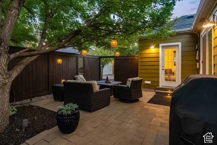 Paved patio space featuring a privacy fence, mature trees, exterior wall lighting, and a white paneled door