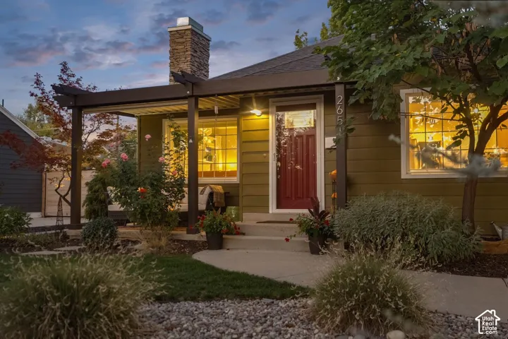 Exterior featuring a stacked stone chimney, covered porch with a pergola, red panel door, and established landscaping with shrubbery and trees