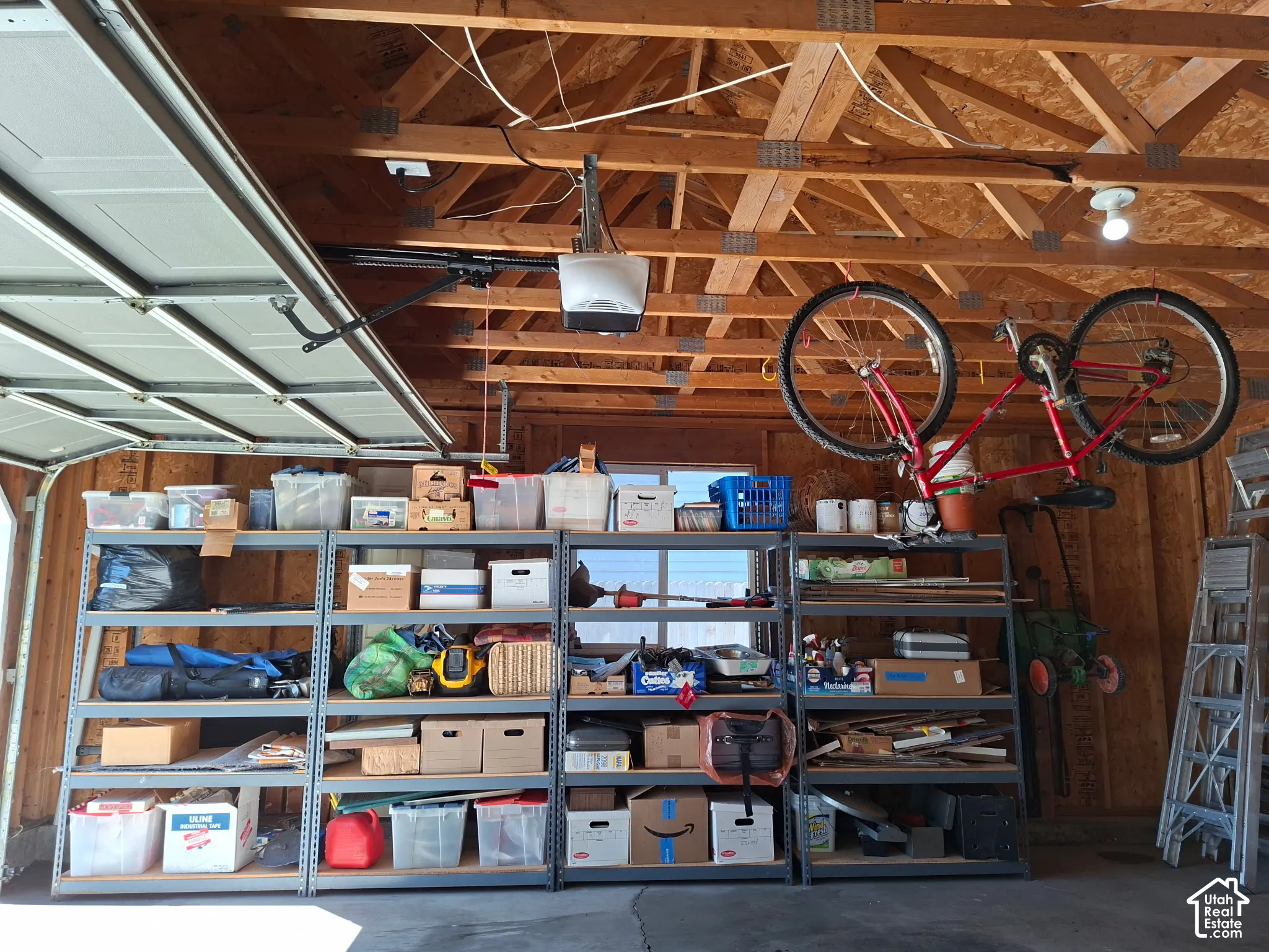 Exposed wood ceiling joists, overhead garage door with automatic opener, built-in shelving units, concrete flooring, and wall-mounted lighting fixture