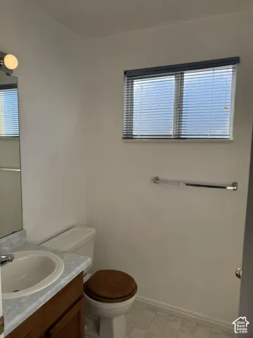 White wall finishes, window with blinds, vanity with integrated sink, wood-finish cabinet, and a white toilet