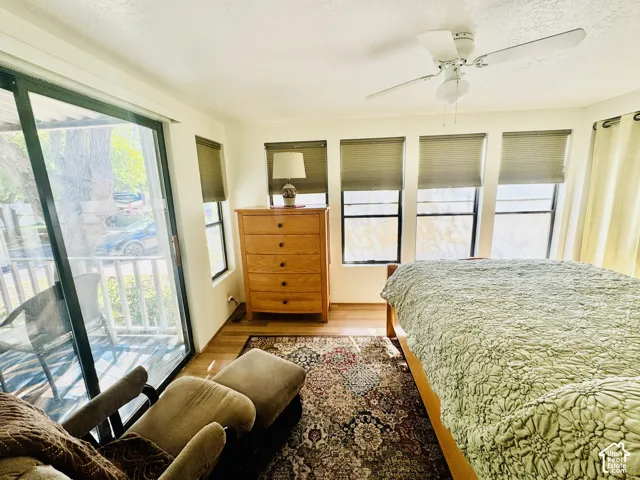 Sliding glass door access to a balcony, wood-finish flooring, ceiling fan with light fixture, and multiple windows with blinds