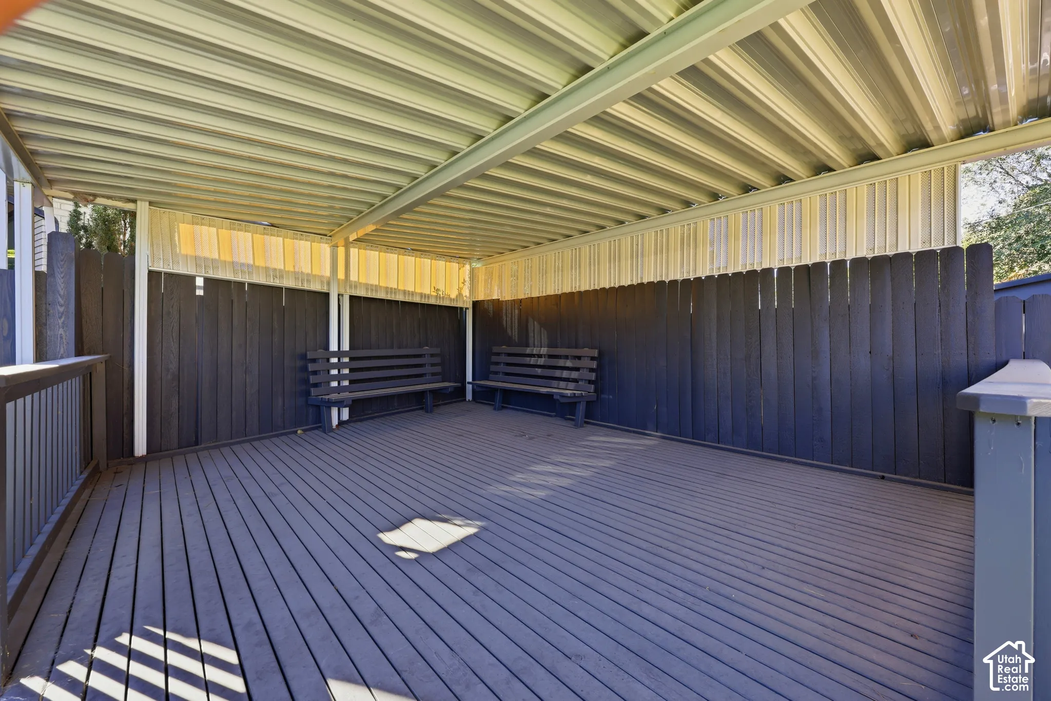 Covered outdoor deck with slatted wood flooring, enclosed by a privacy fence, and featuring a corrugated metal roof