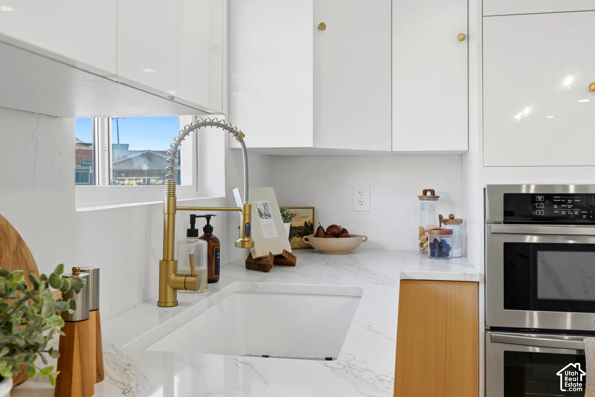 White upper cabinetry with brass-finish hardware, built-in stainless steel appliances, white countertop with veining, single-basin sink, and brass-finish faucet