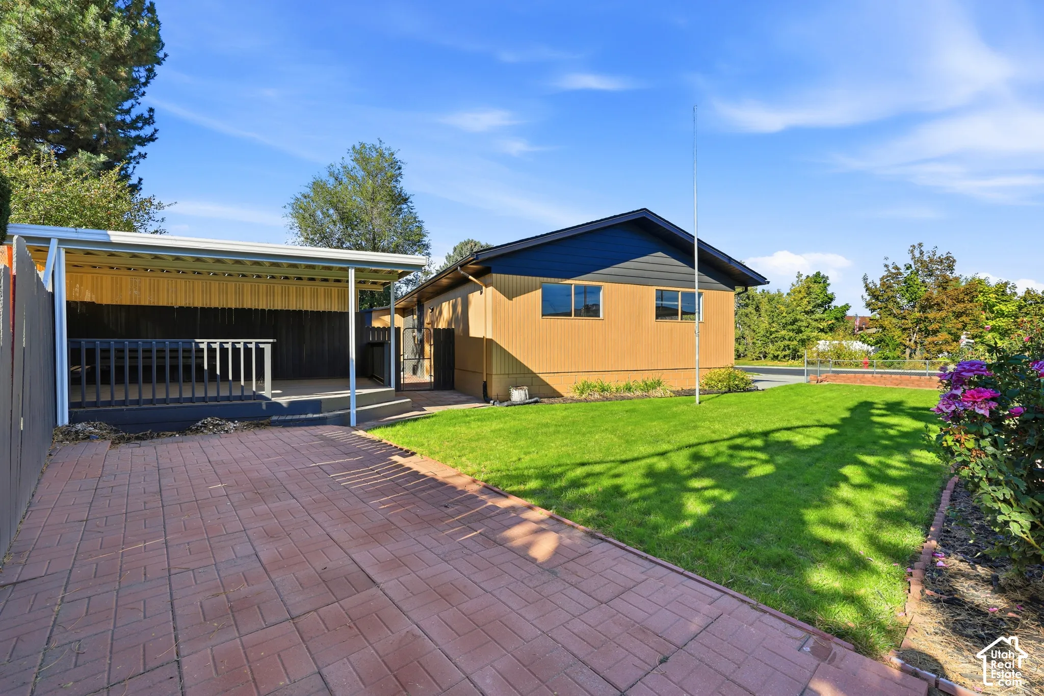 Covered patio with railing, paver driveway, expansive lawn, and landscaping with shrubs