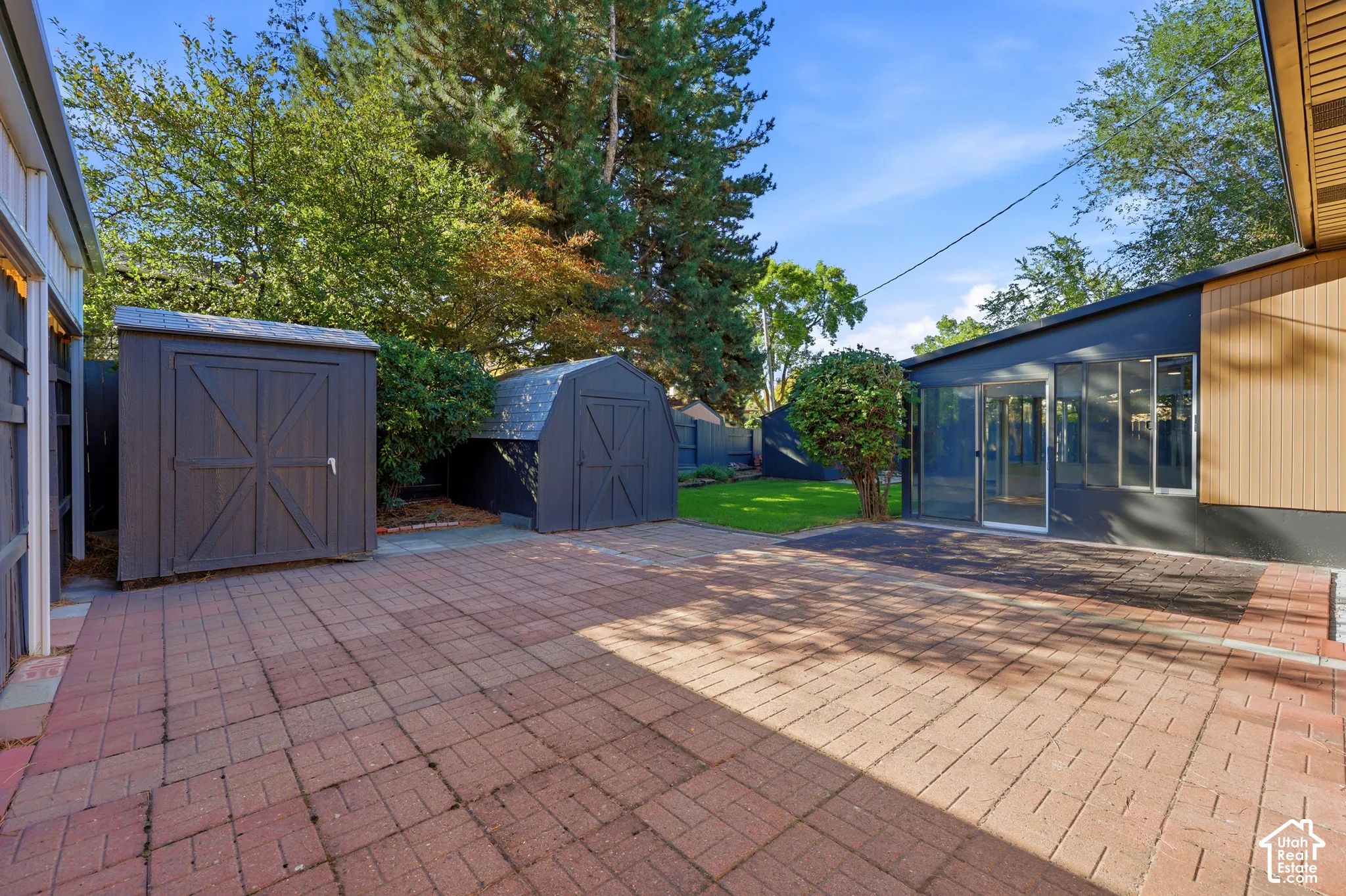 Expansive brick paver patio with two storage sheds, manicured lawn, and mature trees