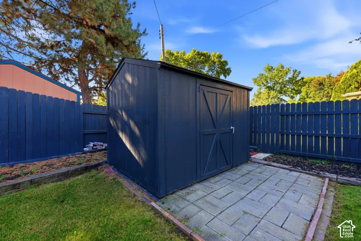 Exterior storage shed with a dark finish, framed by a wood picket fence, a paver patio base, and a grass lawn