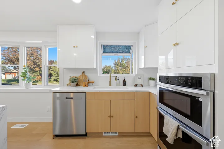 Modern kitchen featuring white upper cabinetry, wood-finish lower cabinetry, integrated stainless steel appliances, a bay window, and a single basin sink with a gold-finish faucet