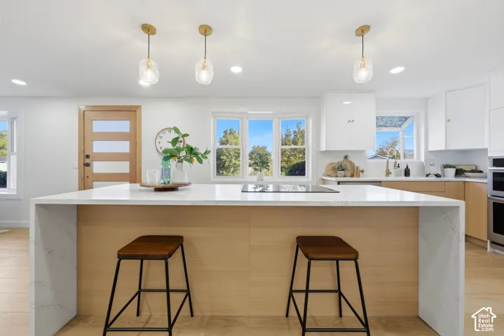 Spacious kitchen featuring an extended island with a waterfall countertop, integrated cooktop, brass pendant lighting, light wood-finish cabinetry, and white upper cabinetry