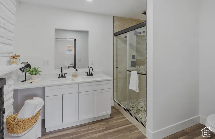 Dual vanity with white cabinetry and quartz-like countertop, matte black fixtures, glass-enclosed shower with tiled walls and pebble flooring, and wood-finish flooring
