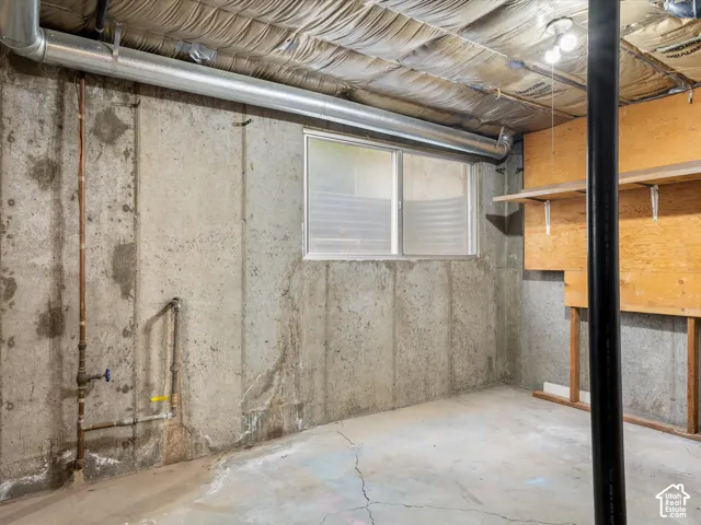 Unfinished utility space featuring exposed concrete walls, concrete flooring, sliding window with a window well, and built-in wooden shelving
