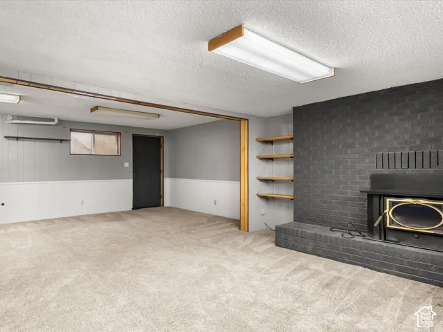 Painted brick fireplace surround featuring an inset wood-burning stove, wall-mounted wood shelving, neutral carpeting, and a textured ceiling
