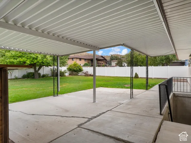 Covered concrete patio featuring metal support columns, black metal railings, and an expansive lawn enclosed by white vinyl fencing