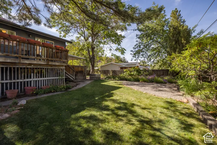 Tiered wooden deck, lower-level covered patio, manicured lawn, established landscaping, and gravel parking area