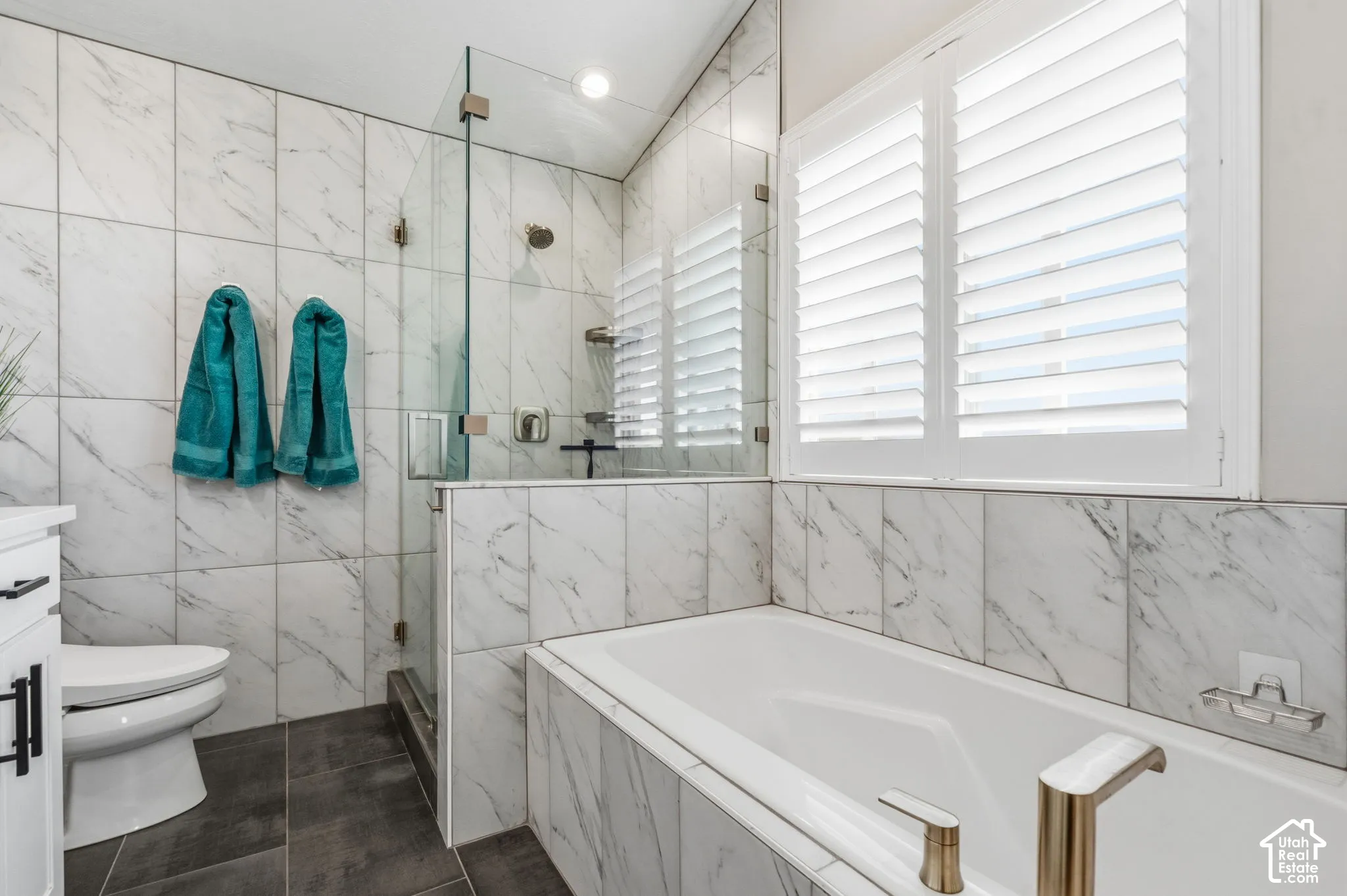 Walk-in shower with glass enclosure, white rectangular soaking tub with gold-tone fixtures, white plantation shutters, and large-format tile walls