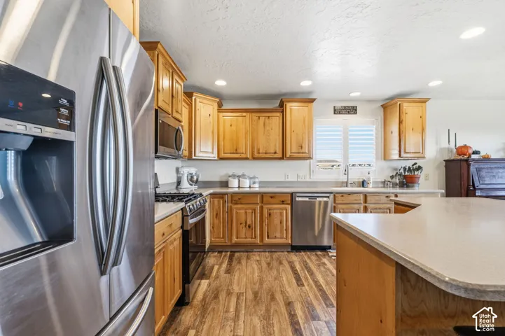 Stainless steel appliances, wood-finish flooring, light wood cabinetry, recessed lighting, and a kitchen island with a light-toned countertop