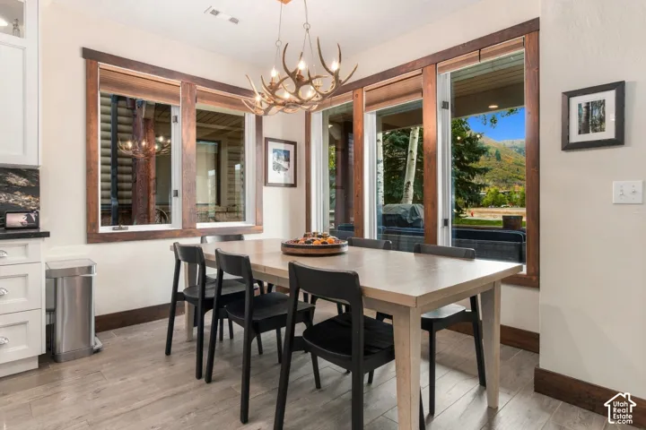 Spacious dining area featuring wood-finish flooring, a contemporary antler-inspired chandelier, white cabinetry with dark countertops, and large windows with wood trim