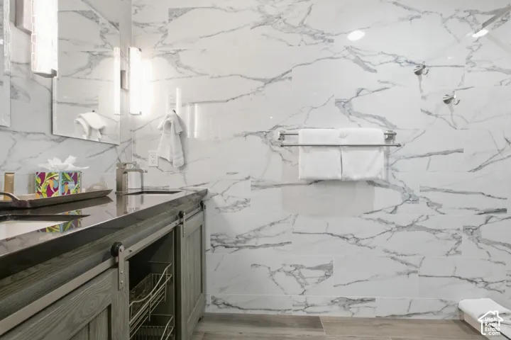 White large-format wall tiles with natural veining, gray wood-finish vanity with sliding barn door hardware, undermount sink, dark countertop, and brushed nickel fixtures