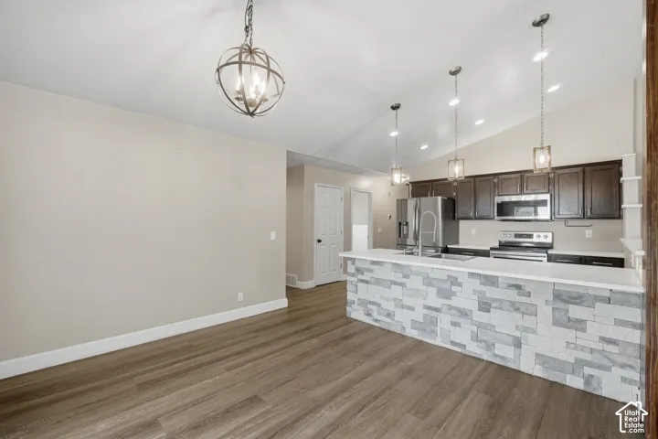 Vaulted ceiling, wood-finish flooring, kitchen island with stone facade, dark wood cabinetry, and stainless steel appliances