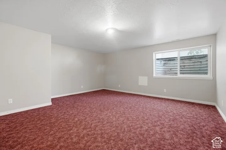 Spacious room featuring neutral painted walls, textured ceiling, a window with a window well, and red patterned carpeting