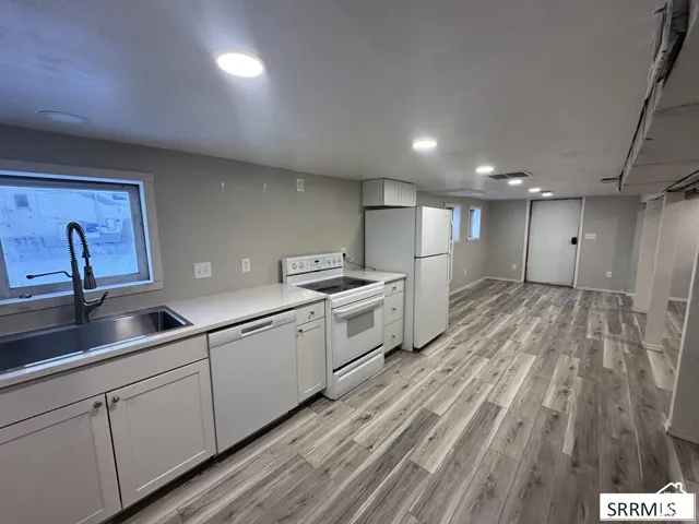 Kitchen area featuring white cabinetry, a stainless steel sink with a gooseneck faucet, white appliances, light-toned countertops, and wood-finish flooring