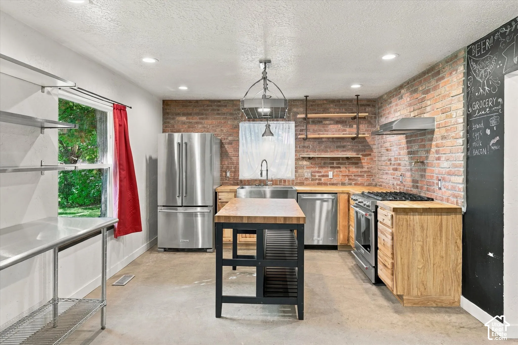 Modern kitchen featuring stainless steel French-door refrigerator and range, wood-finish cabinetry, butcher-block countertops, exposed brick accent wall, pendant lighting, and a central island with black metal base and wood-finish top