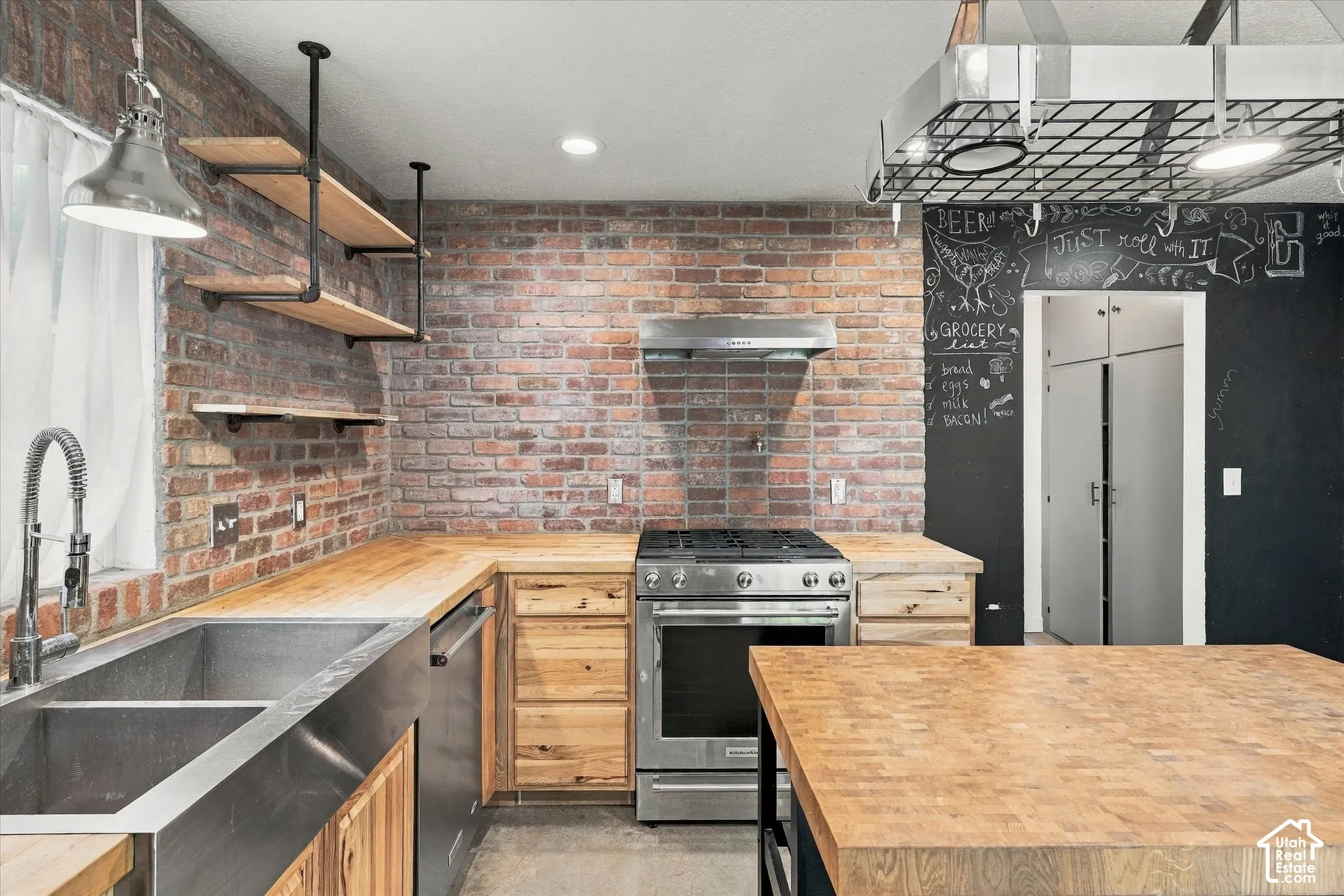 Industrial-modern kitchen featuring exposed brick walls, stainless steel range with hood, wood cabinetry, butcher block countertops, undermount sink with gooseneck faucet, chalkboard accent wall, pendant lighting, and stainless steel refrigerator