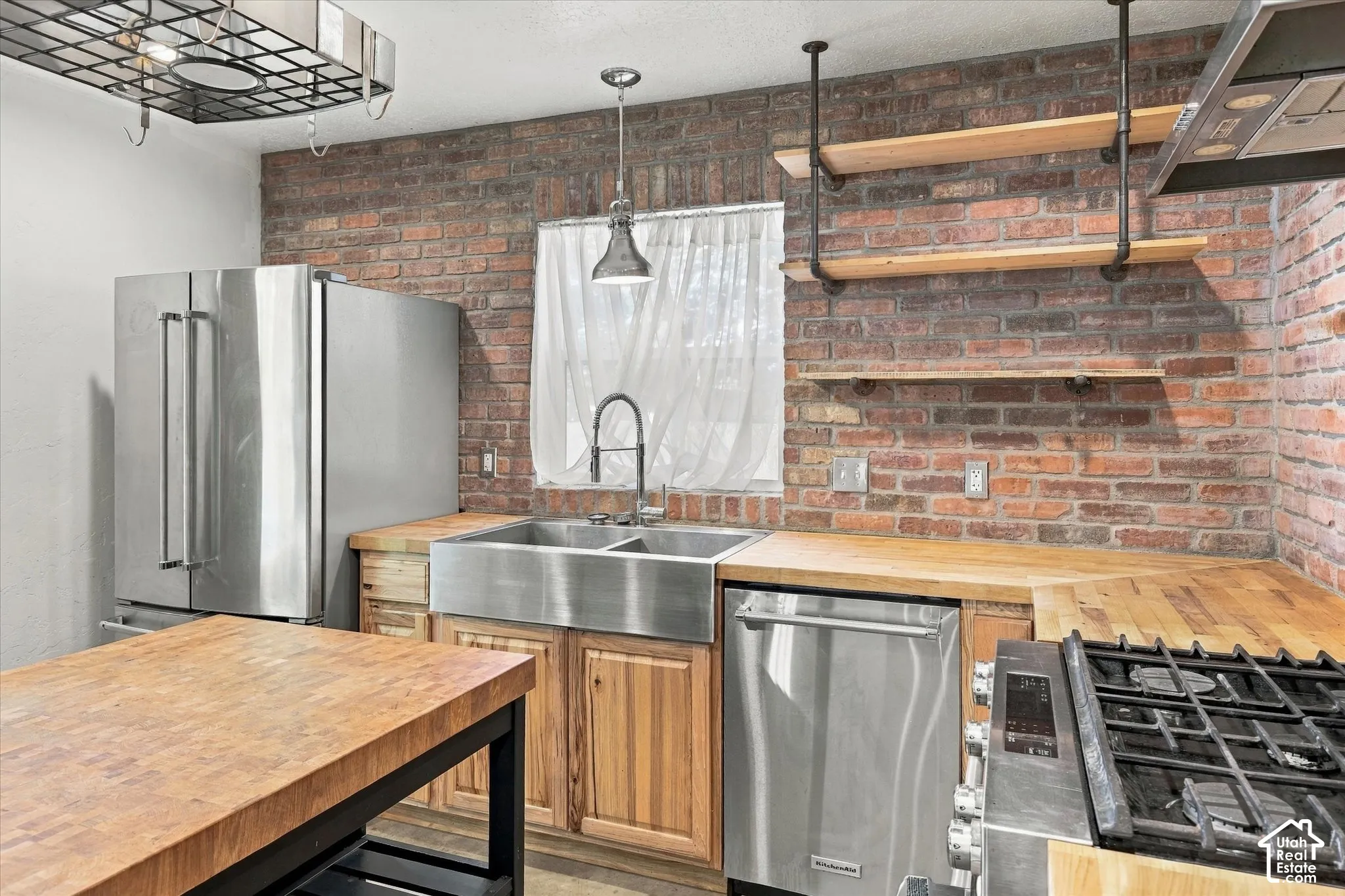 Exposed brick accent wall, stainless steel appliances, butcher block countertops, farmhouse sink, and open shelving