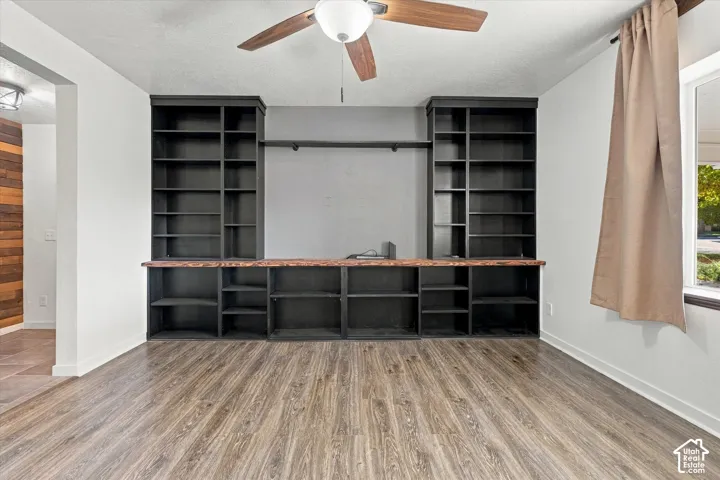 Spacious bonus room featuring extensive built-in black shelving across the entire wall, wood-finish flooring, ceiling fan, and abundant natural light from dual windows