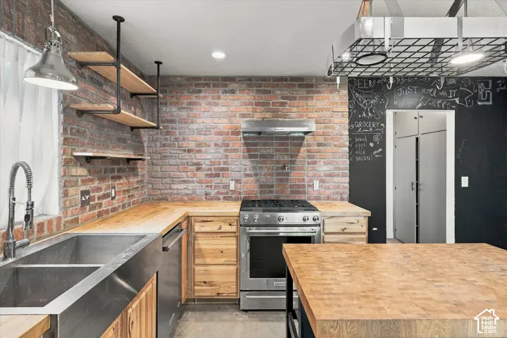 Industrial-modern kitchen featuring exposed brick walls, stainless steel range with hood, wood cabinetry, butcher block countertops, undermount sink with gooseneck faucet, chalkboard accent wall, pendant lighting, and stainless steel refrigerator