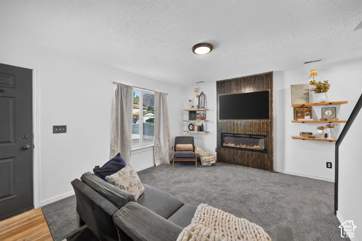 Living area featuring a floor-to-ceiling accent wall, built-in electric fireplace, wall-mounted display shelving, a large window, and gray carpeting