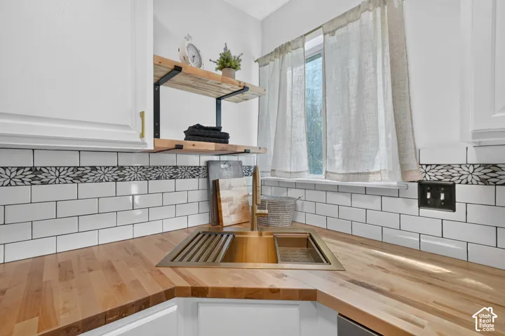 Solid wood countertops, white subway tile backsplash with decorative accent, undermount sink with integrated drying rack, white cabinetry, and open shelving