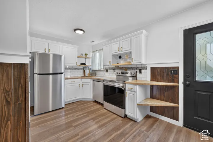 White cabinetry with gold-tone hardware, wood-finish flooring, stainless steel appliances, subway tile backsplash, and corner shelving