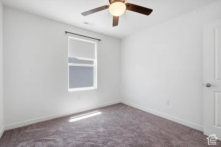 White walls with grey carpeting, featuring a window with a roller shade, a ceiling fan with integrated lighting, and a white paneled door