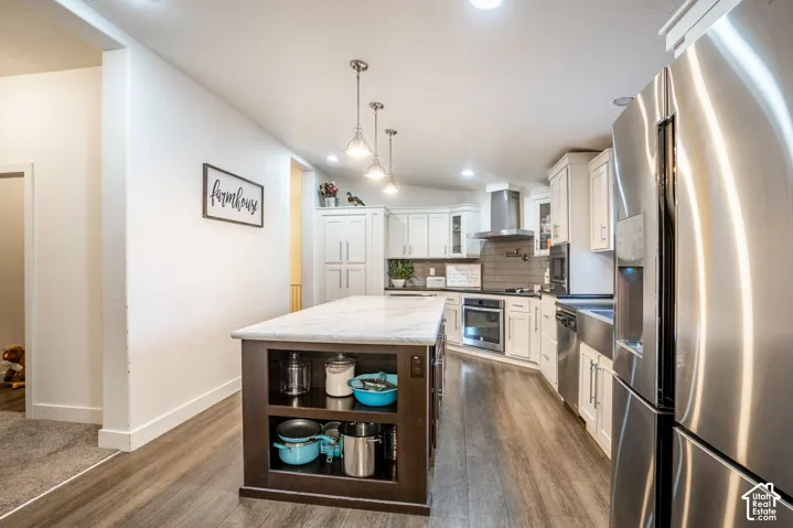 Modern kitchen featuring a large center island with wood-finish cabinetry and open shelving, stainless steel appliances including a refrigerator and range hood, white shaker-style upper cabinetry, pendant lighting, and wood-finish flooring throughout the
