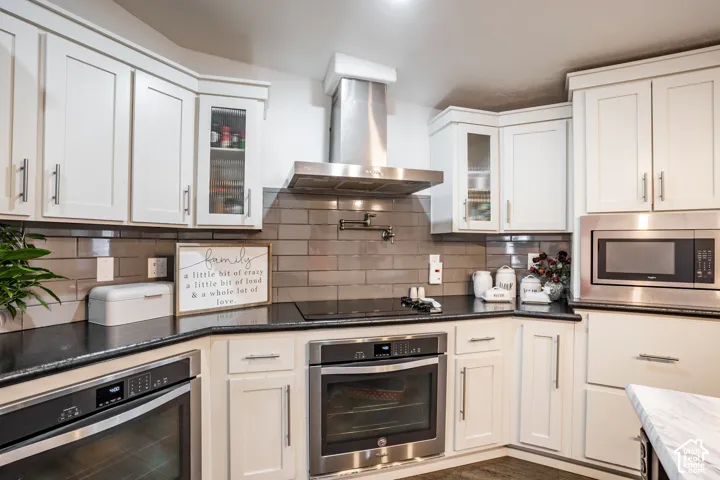 Elegant kitchen featuring white shaker-style cabinetry with brushed nickel hardware, stainless steel appliances including a dual oven configuration, subway tile backsplash, dark stone countertops, and a stainless steel range hood with integrated lighting