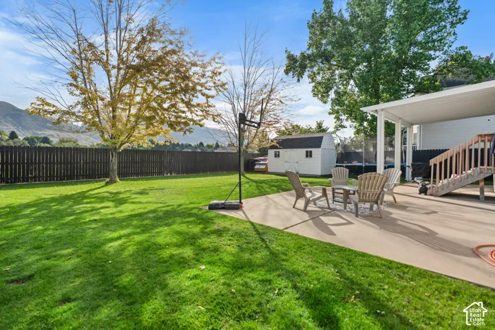 Expansive backyard featuring a manicured lawn, full privacy fence, concrete patio, covered outdoor living space, and a storage shed