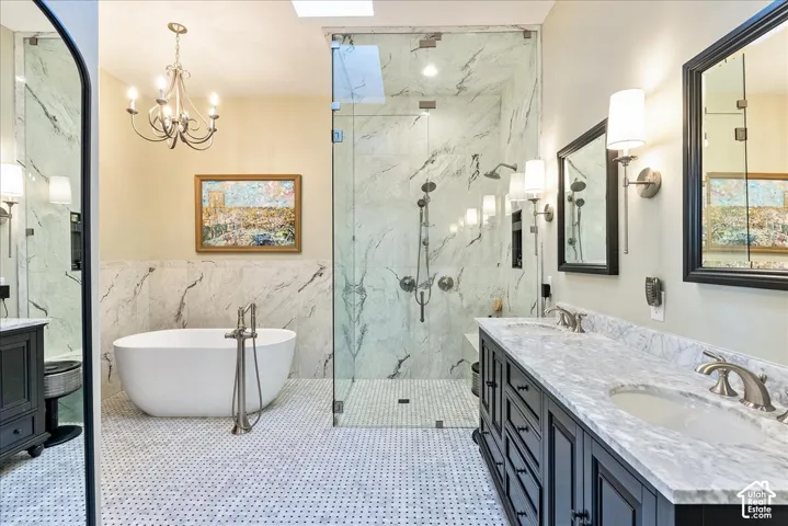 Freestanding soaking tub with floor-mounted faucet, dual vanity with undermount sinks and stone countertops, glass-enclosed shower with multiple showerheads, and mosaic tile flooring