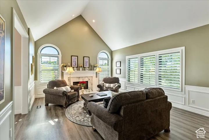 Vaulted ceiling, wood-finish flooring, white wainscoting, fireplace with a marble-look surround, and white plantation shutters