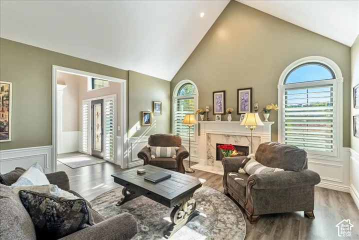 Vaulted ceiling, arched windows with plantation shutters, wood-finish flooring, white fireplace with marble-look surround, and wainscoting