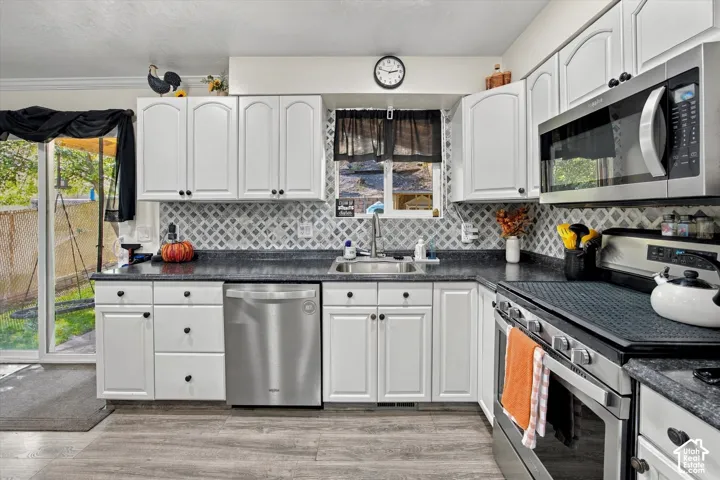White cabinetry with black hardware, stainless steel appliances, patterned tile backsplash, dark countertops, and wood-finish flooring