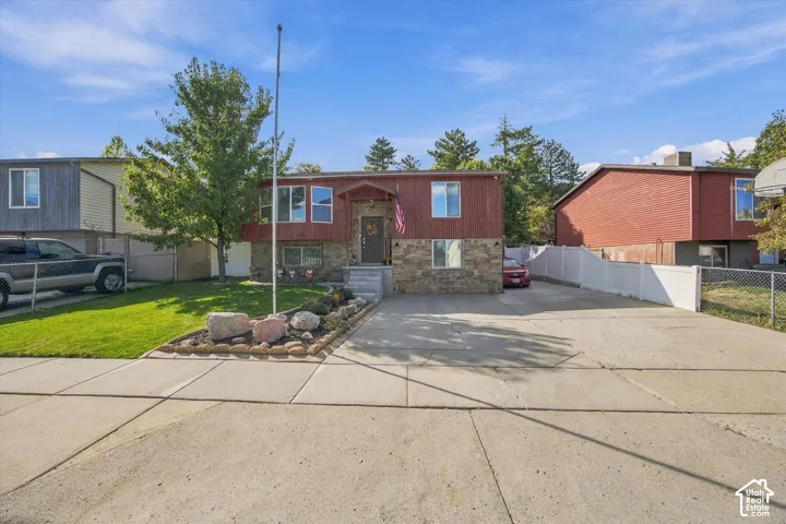 Split-level residence featuring a stone and siding exterior, concrete driveway, front yard with mature landscaping, and a white privacy fence