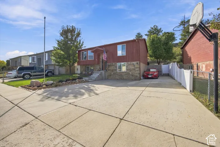 Multi-level residence featuring a stone and wood-panel exterior, a concrete driveway, a front yard with established landscaping, and a side yard with a white privacy fence