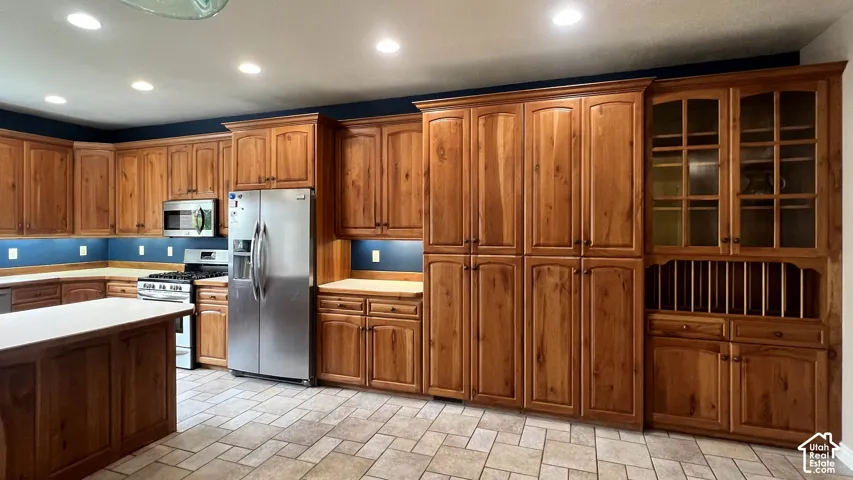 Expansive kitchen featuring wood cabinetry, stainless steel appliances, a center island with a light-toned countertop, and tiled flooring