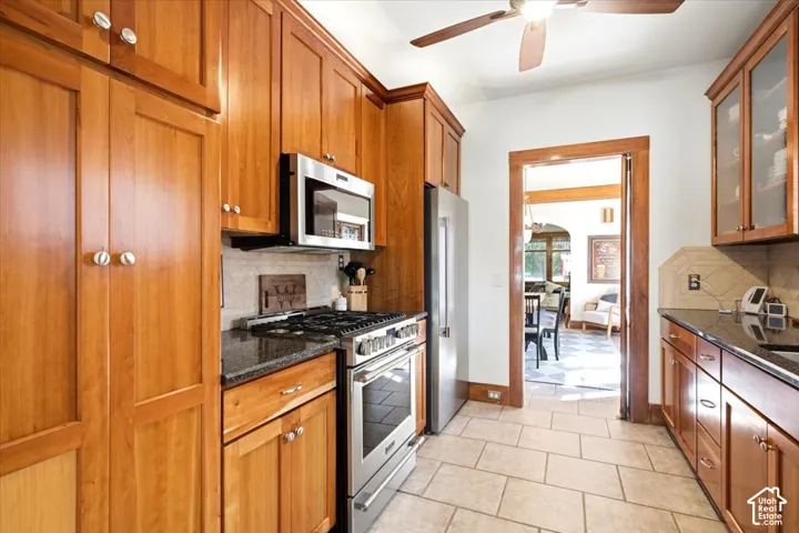 Kitchen featuring wood-finish cabinetry, stainless steel appliances, dark countertops, and light tile flooring