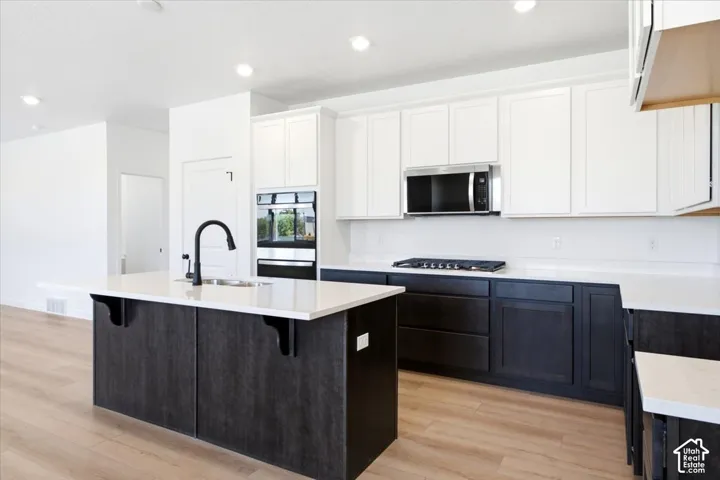 Kitchen featuring white upper cabinetry, dark lower cabinetry, a center island with a sink, stainless steel appliances, and light wood-finish flooring