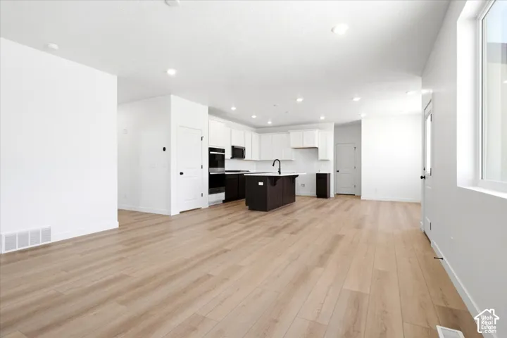 Open-concept layout featuring wood-finish flooring, recessed lighting, a kitchen island with a black faucet, white shaker cabinetry, and built-in double ovens