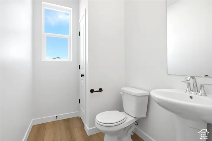 Pedestal sink with chrome faucet, toilet, wall mirror, window, and wood-finish flooring