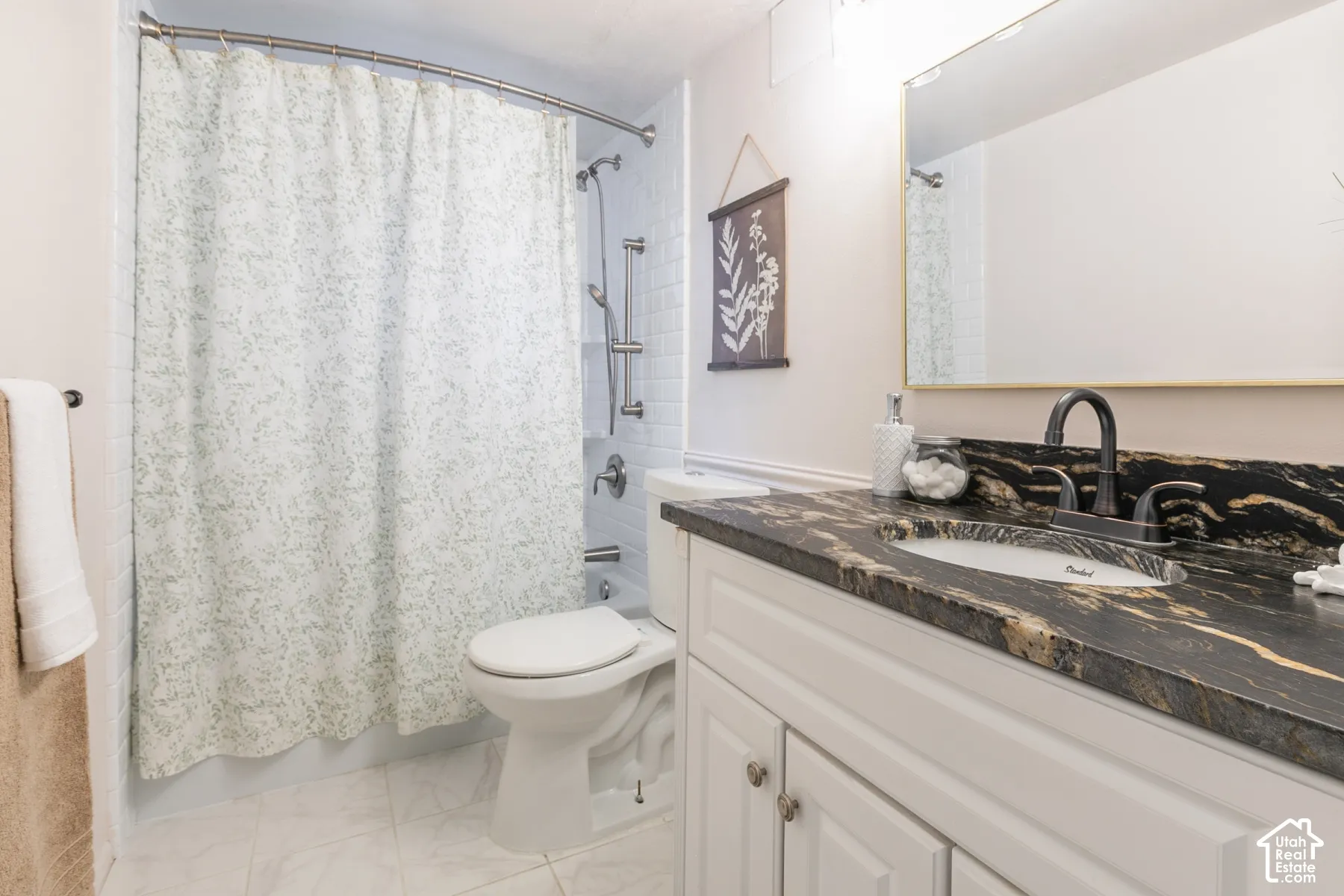 Bathroom featuring a white vanity with paneling, a dark countertop with gold veining, a wall-mounted mirror, a tiled shower with a handheld showerhead, and white floor tiles