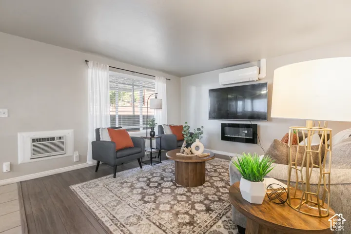 Wood-finish flooring, wall-mounted air conditioning unit, built-in electric fireplace, and a large window with blinds