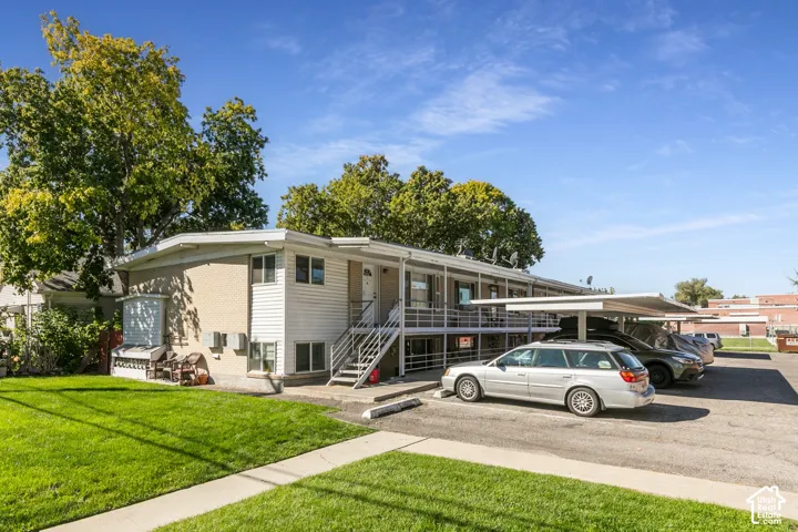 Two-story building exterior featuring brick and siding finishes, multiple balconies, exterior staircases, and an expansive paved parking area with covered carports